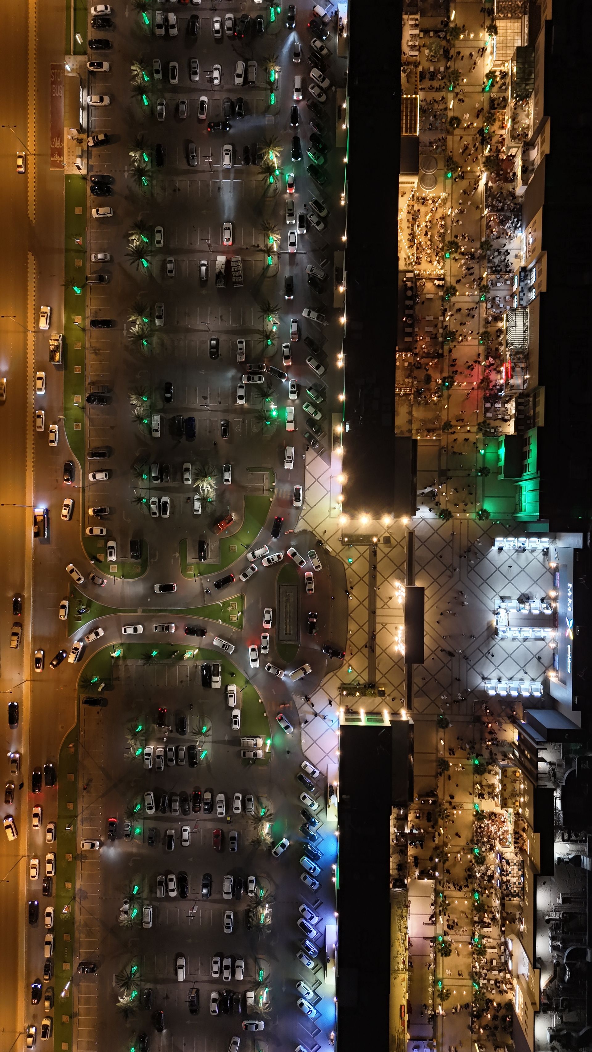 Aerial view at night of a Saudi city plaza: crowded terraces and walkways beside rows of parked cars and traffic